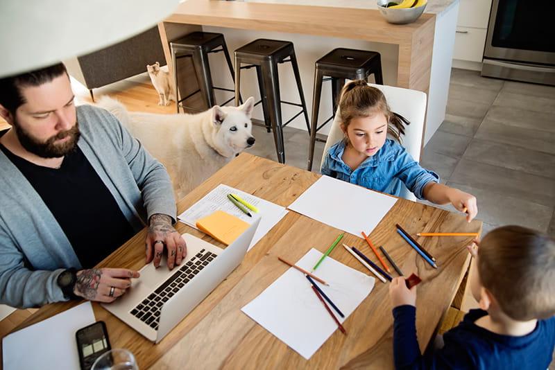 father uses laptop in kitchen while kids draw dog